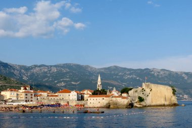 Eski Budva kasabasının kalesi denize doğru itildi. Kale duvarlarının altında yüzen turistlerin olduğu sahil ve iskele.