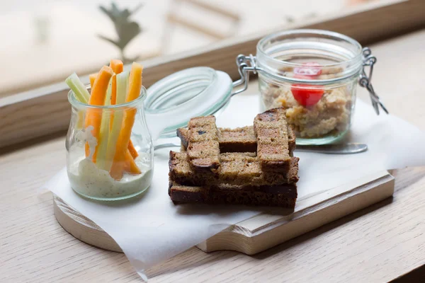 Hummus y pan sobre tabla de madera. Zanahorias en un frasco de vidrio — Foto de Stock