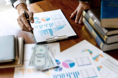 İş adamları masadaki grafiği göz önünde bulundurarak çalışıyorlar. Masada para ve kitaplar var.