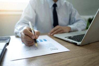 Masaya evrak yazmak için kalem tutan bir işadamı CEO 'su masadaki dizüstü bilgisayarla bir rapor hazırlayacak..