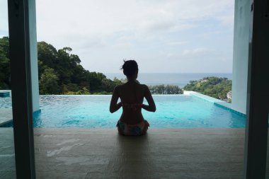 Yüzme havuzunun kenarında oturan ve deniz manzarasına bakan, bikinili zayıf genç bir kadın..