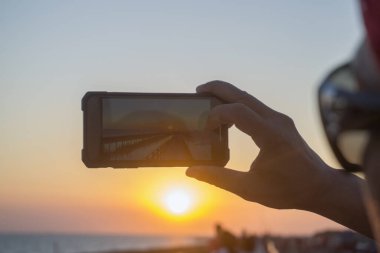 Gün batımının fotoğraflarını çekmek için elinde cep telefonu tutan bir adam..