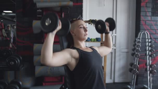 Mulher muscular senta-se no banco e levanta halteres no ginásio — Vídeo de Stock