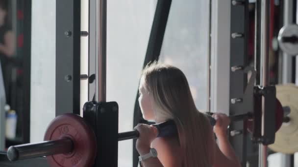 Rückansicht einer blonden Frau macht Kniebeugen mit Langhantel an Schmied-Gerät im Fitnessstudio — Stockvideo