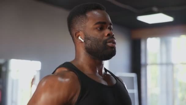 Close up de Africano americano homem fisiculturista com fones de ouvido sem fio realizando Side raise exercício com halteres no ginásio — Vídeo de Stock