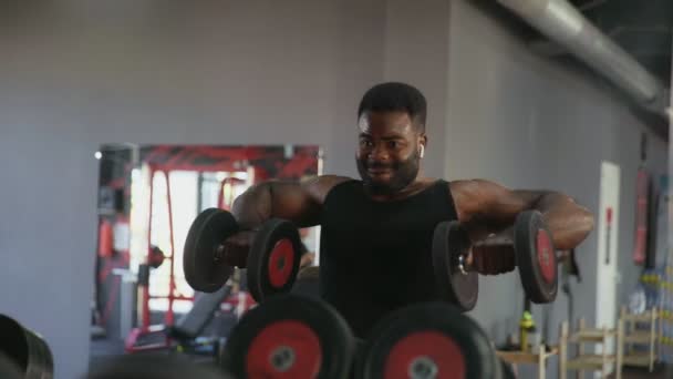 Uomo afroamericano bodybuilder con cuffie wireless esecuzione Side sollevare esercizio con manubri e si guarda allo specchio in palestra — Video Stock