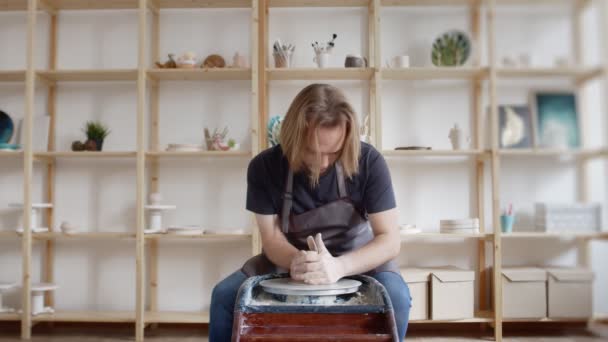 Молодой мужчина-ремесленник, сидящий на стуле и создающий глиняный сосуд на колесах во время работы в просторной профессиональной керамике — стоковое видео