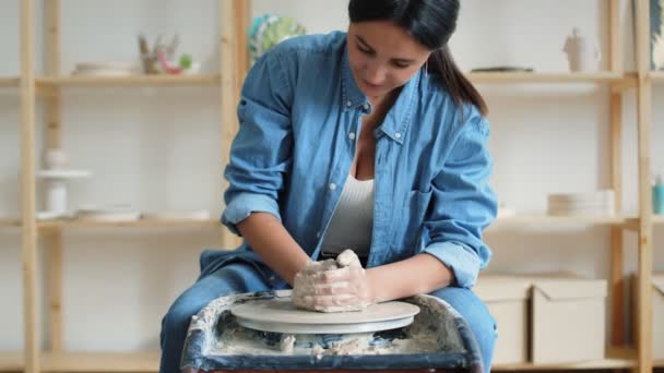 Женщина-ремесленница, сидящая на стуле и создающая глиняный сосуд на колесе во время работы в просторной профессиональной керамике — стоковое видео