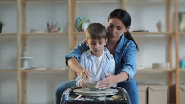 Женщина ремесленник и ее сын сидят на стуле и изготовление глины вместе сосуд на колесе во время работы в просторной профессиональной керамики — стоковое видео