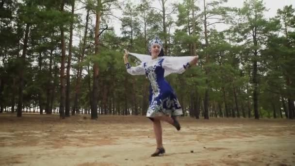 Красивая женщина в голубом русском традиционном народном костюме с платком танцует в лесу — стоковое видео