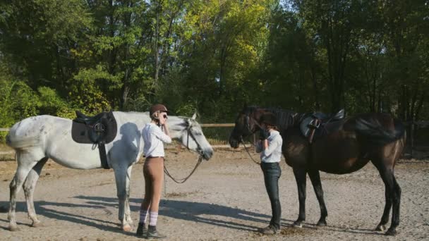 Deux cavalières en vêtements d'équitation et casques se préparent à monter sur leurs chevaux — Video