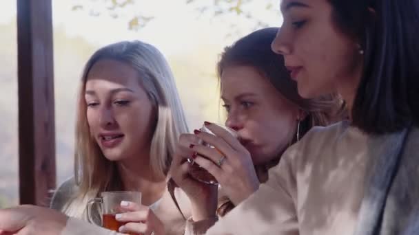Tre glada kvinnor med en filt sittande på sina axlar i terrass och dricka te — Stockvideo