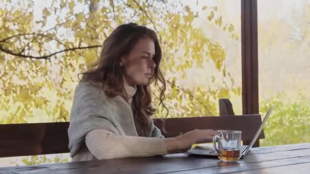 Mujer joven con manta en los hombros utiliza portátil y bebe té en la terraza al aire libre — Vídeos de Stock