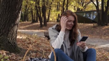 Güzel genç bir kadın parkta battaniyeye oturur ve güneşli bir sonbaharda telefonu kullanır.