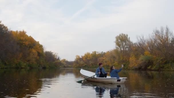 Jovem cara bonito e uma linda garota tira uma foto em um barco em um lago em uma floresta de outono fada — Vídeo de Stock