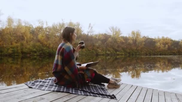 Junge schöne Frau mit einer Decke auf den Schultern liest Buch und trinkt Kaffee, während sie am Fluss stits po piea — Stockvideo