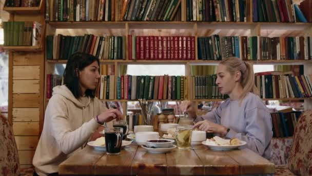 Arka planda kitap rafı olan iki genç kadın kafede oturuyor. — Stok video