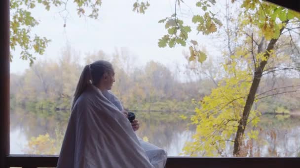 Jonge blonde vrouw met deken op de schouders drinkt warme thee en zit op terras bij de rivier — Stockvideo