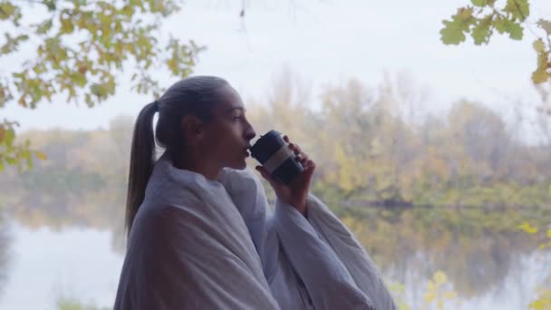 Blisko blondynka z kocem na ramionach pije gorącą herbatę i siedzi na tarasie nad rzeką — Wideo stockowe