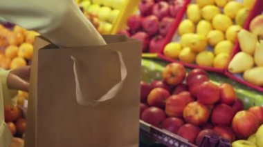 Sarı ceketli bir kadın markette kırmızı elmaları bir poşete koyuyor.