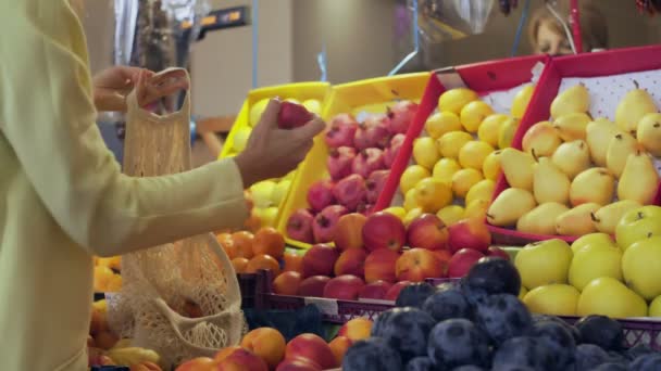 Achteraanzicht van de vrouw in gele jas zet rode appels in string zak op de markt — Stockvideo