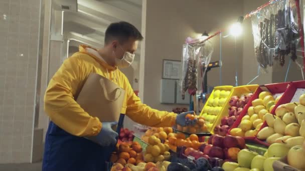 Een man met beschermende masker en handschoenen kiest een verse mandarijnen, zet ze in een papieren zak en geeft het aan de verkoper op de markt — Stockvideo