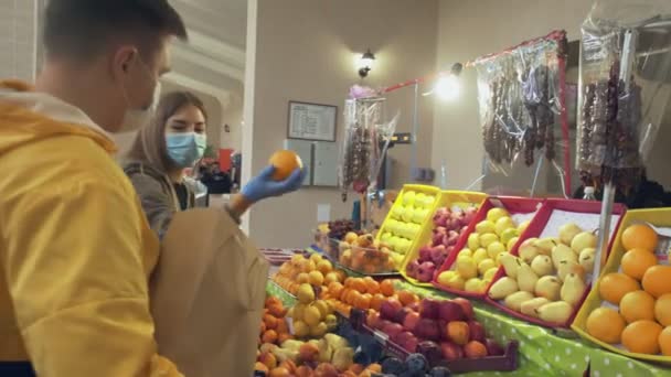 Jonge paar met beschermende masker en handschoenen kiest een verse sinaasappels, zet ze in een papieren zak ang geeft het aan verkoper in de markt — Stockvideo