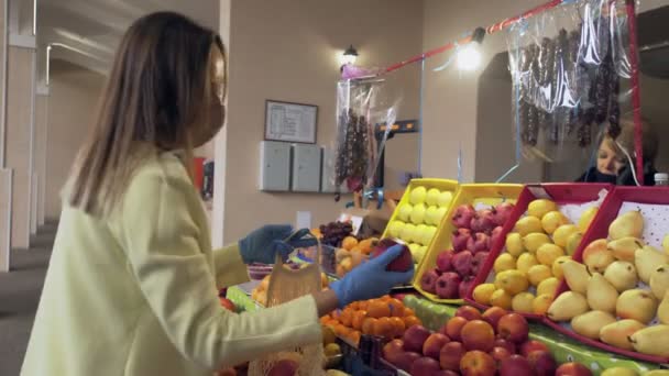 Donna in cappotto giallo, maschera protettiva e guanti blu sceglie una mela fresca al mercato — Video Stock