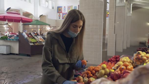 Молодая женщина в защитной маске и синих перчатках нюхает и выбирает персики на полках магазинов на рынке — стоковое видео