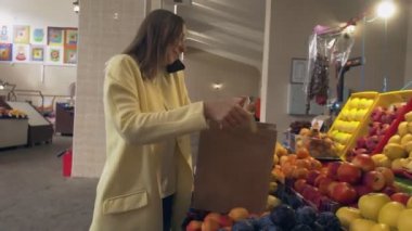 Sarı ceketli kadın telefonla konuşuyor ve kağıt torbaya taze kırmızı elmalar koyuyor.