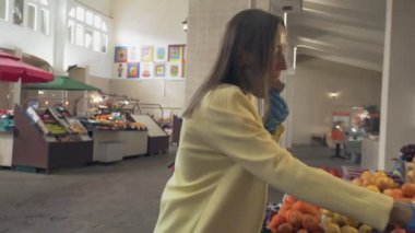 Sarı ceketli, koruyucu maskeli ve mavi eldivenli bir kadın marketteki taze meyveleri koklar ve seçer.
