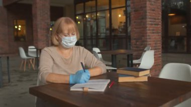 Koruyucu maskeli ve eldivenli kadın kitap yazarı kafede çalışıyor.