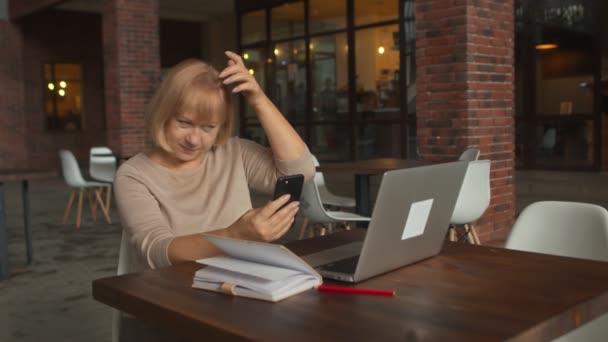 Lady olha para o telefone e cabelo correto, enquanto se senta no café com notebook — Vídeo de Stock