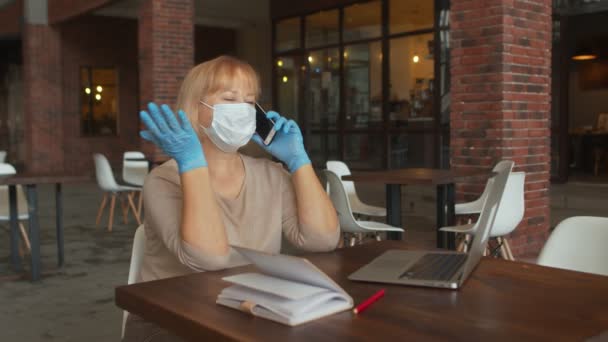 Señora con máscara protectora y guantes habla por teléfono mientras se sienta en la cafetería con portátil — Vídeos de Stock