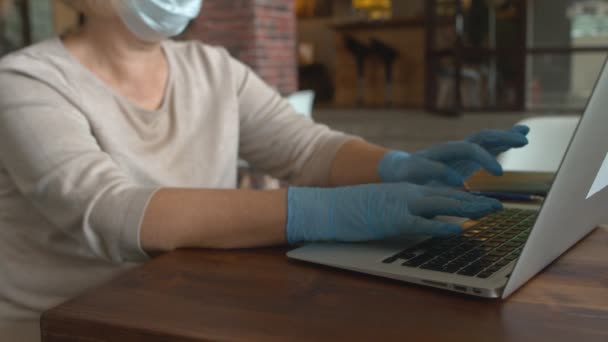 Zblízka dáma s ochrannou maskou a rukavicemi pracuje na notebooku v kavárně — Stock video