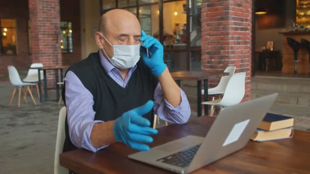 Hombre de negocios senior con máscara protectora y guantes habla por teléfono y trabaja en el portátil en la cafetería — Vídeos de Stock