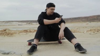 Genç bir adam kaykayın üzerinde oturuyor ve çölde telefon kullanıyor.