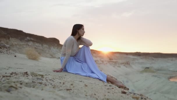 Vacker ung kvinna i blå kjol sitter på toppen av sand kulle med solnedgång på bakgrunden — Stockvideo