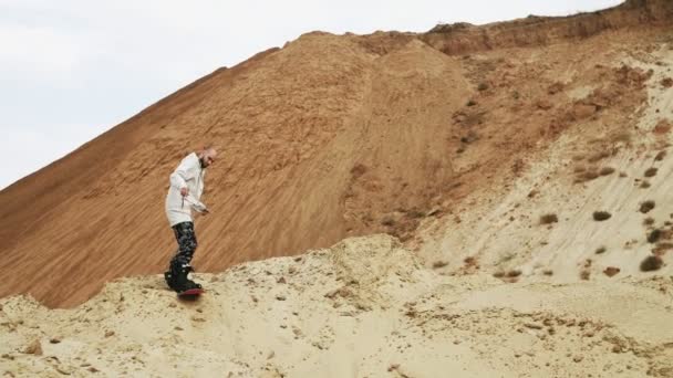 Hombre guapo sandboards una montaña en desierto o cantera de arena — Vídeos de Stock