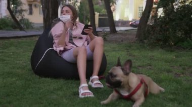 Güzel genç bir kadın Fransız bulldoguyla fasulye koltukta oturuyor ve parkta telefon kullanıyor.