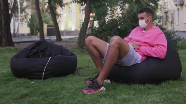 Młody mężczyzna i kobieta w maskach ochronnych siedzą w torbach w parku i używają gadżetów — Wideo stockowe