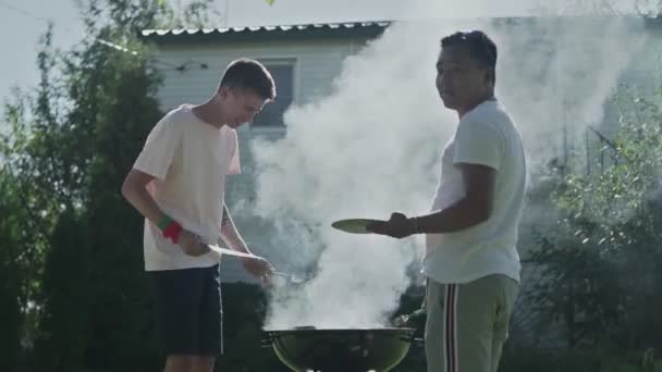 Two happy men cooking grilling bacon and sausages on grid — Stock Video