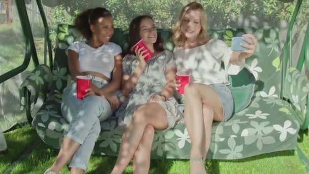 Három boldog fiatal nő piros poharakkal ül a hintán és szelfit készít. — Stock videók
