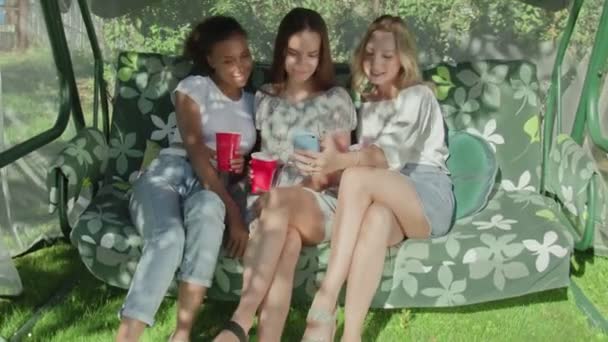 Three happy young women with red cups sits on swing and looks at phone — Stock Video