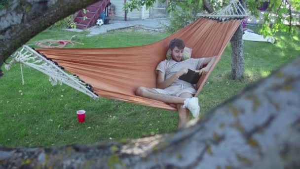 Un giovane che legge il libro mentre si trova in un hammock nel cortile posteriore — Video Stock