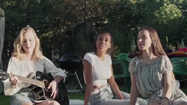 Γυναίκα παίζει κιθάρα και δύο γυναίκες τραγουδούν. Οι φίλες κάθονται σε μια κουβέρτα στο πάρκο. — Αρχείο Βίντεο