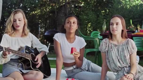 Νεαρή γυναίκα παίζει κιθάρα και δύο γυναίκες τραγουδούν και τρώνε καρπούζι. Οι φίλες κάθονται σε μια κουβέρτα στο πάρκο. — Αρχείο Βίντεο