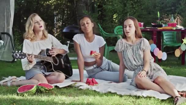 Young woman playing at the guitar and two woman are singing and eating watermelon. Girlfriends sit on a blanket in park — Stock Video