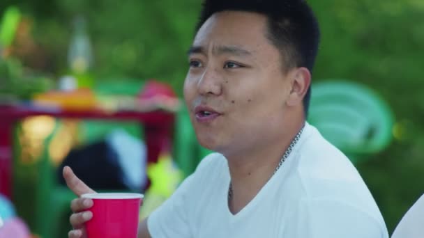 Lähellä aasialainen mies laulaa ystäviensä kanssa. Ystävät istuvat huovan päällä puistossa. — kuvapankkivideo