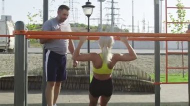 Kıdemli antrenör, genç bayan sporcusunu yatay çizgide kendini yukarı çekmesi için eğitiyor. Açık havada..